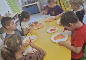 Grupa dzieci wykonuje dynie z wykorzystaniem talerzyka i farby pomarańczowej przy stolikach