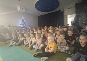 Dzieci siedzą na dywanie i oglądają koncert